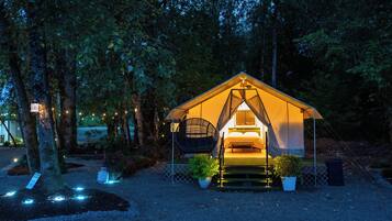 Une tente équipée d’un lit et d’un fauteuil suspendu, entourée d’arbres et illuminée par des guirlandes lumineuses.