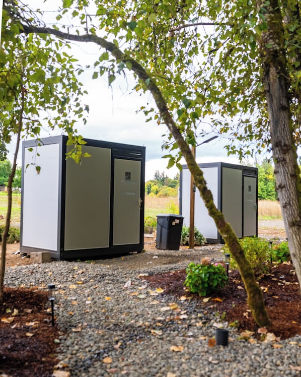 Two large, rectangular, metallic structures with doors, set on a gravel surface, surrounded by trees and greenery.