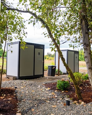 Two large, rectangular, metallic structures with doors, set on a gravel surface, surrounded by trees and greenery. - New Leaf Greenhouse (Cornelius)