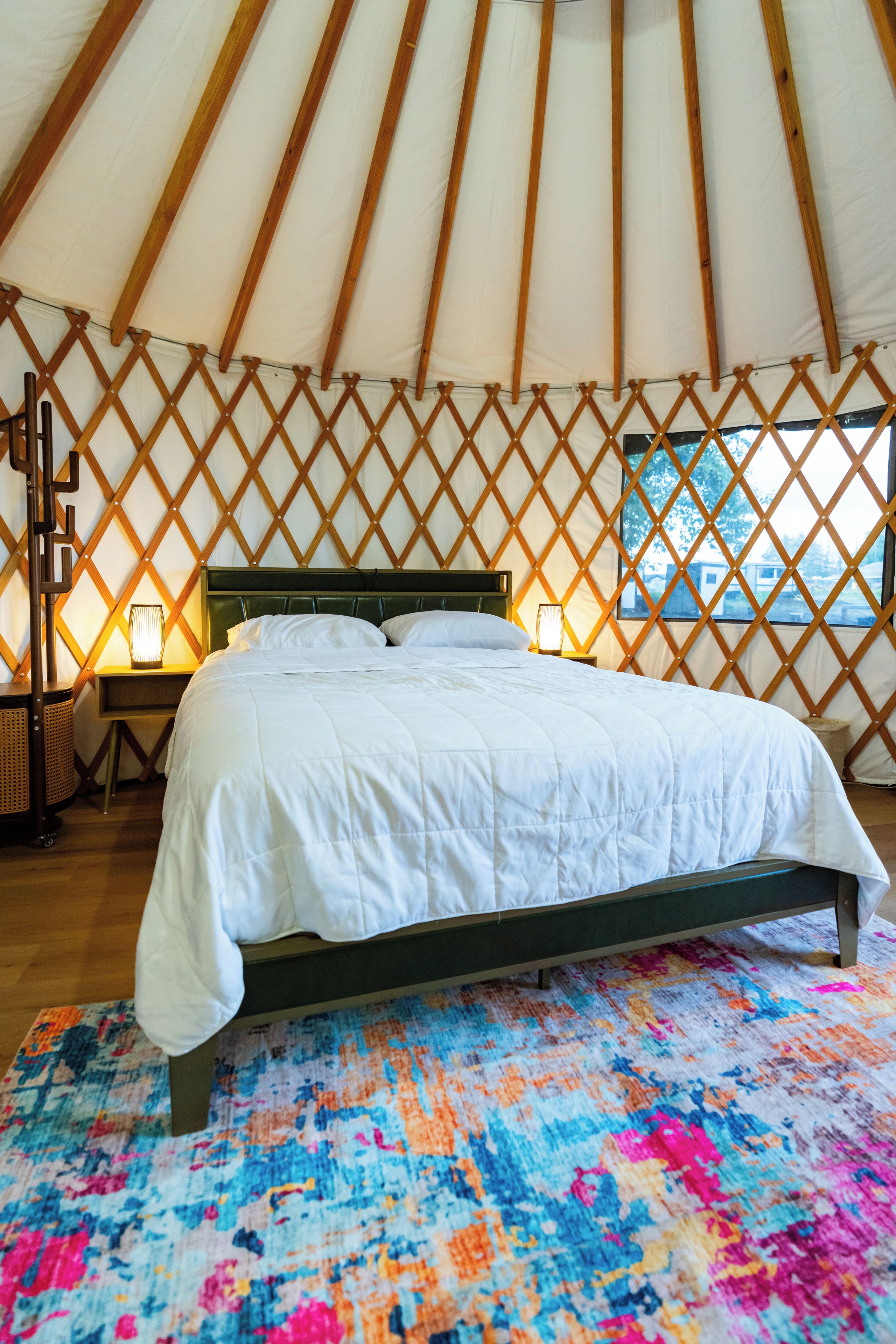 Een bed in een tent met een kleurrijk vloerkleed, houten latenwanden en een nachtkastje met een lamp.