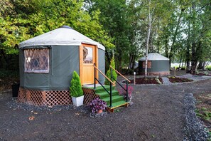 A yurt with a wooden door and a small porch, surrounded by trees and gravel.
