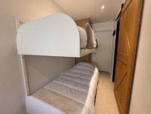 Uma cama beliche com cabeceira branca, uma cama inferior com colchão bege e um guarda-roupa de madeira com porta.