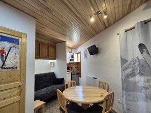 Ein gemütliches Zimmer mit Holzbalkendecke, einem Esstisch mit Stühlen, einer Couch, einer Küchenzeile und einem an der Wand befestigten Fernseher.