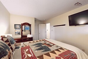 Un dormitorio con cama, cómoda, espejo, televisión y puerta que da a otra habitación.