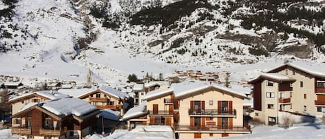 A snow-covered mountain village with chalet-style buildings, a ski lift, and a clear view of the surrounding peaks.