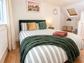 Una cama bien hecha con ropa de cama a rayas y un plaid verde, una lámpara de noche y un cuadro en la pared.