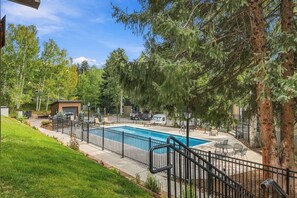 A swimming pool surrounded by a black metal fence, with a small building and outdoor seating area in the background. - Snowlion Vail - Pool, Near Shuttle & Village (Vail)