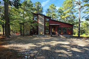 A modern cabin with a stone facade, large windows, and a gravel driveway surrounded by trees. - Happy Hours - Hot Tub, Fire Pit, Coziness (Broken Bow)