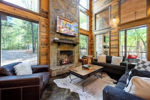 A log cabin interior with a stone fireplace, leather furniture, and large windows offering a view of the forest. - Happy Hours - Hot Tub, Fire Pit, Coziness (Broken Bow)