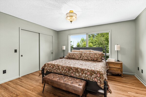 A bedroom with a bed, a bench, a nightstand, a lamp, and a window with a view of trees.