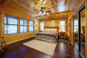 Un dormitorio tipo cabaña de madera con cama, ventilador de techo y ventanas amplias.