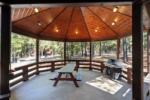 Un área de picnic techada con una mesa de madera y una parrilla, rodeada por una valla de madera y árboles.