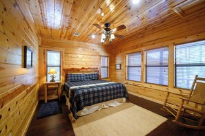 Un dormitorio de cabaña de madera con una cama, un sillón mecedor, una mesita de noche con lámpara y un ventilador de techo.