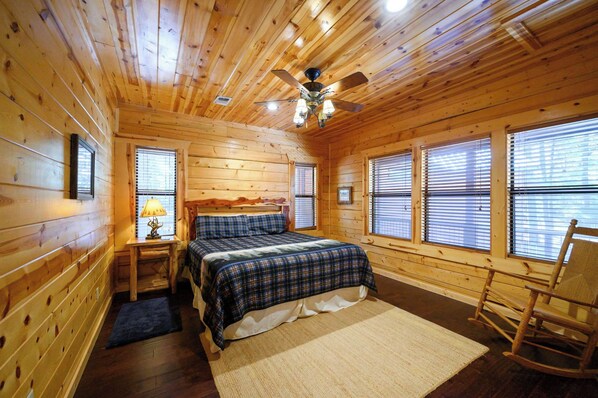 Un dormitorio de cabaña de madera con una cama, un sillón mecedor, una mesita de noche con lámpara y un ventilador de techo.