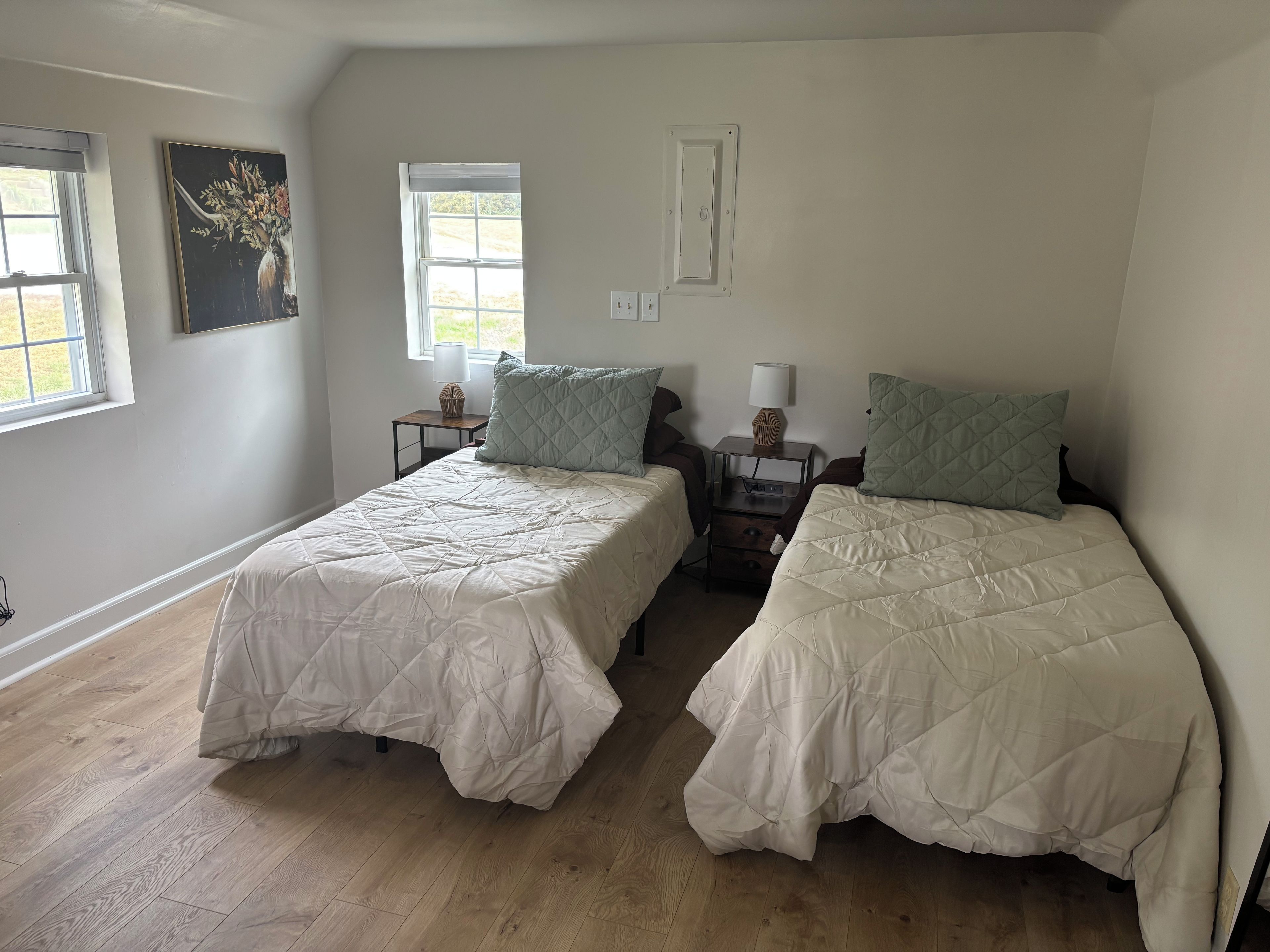 A room with two beds, each with a quilted bedspread and matching pillows, a nightstand with a lamp, a window with a view, and a painting on the wall.