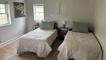 A room with two beds, each with a quilted bedspread and matching pillows, a nightstand with a lamp, a window with a view, and a painting on the wall.