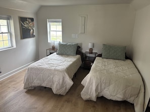 A room with two beds, each with a quilted bedspread and matching pillows, a nightstand with a lamp, a window with a view, and a painting on the wall.