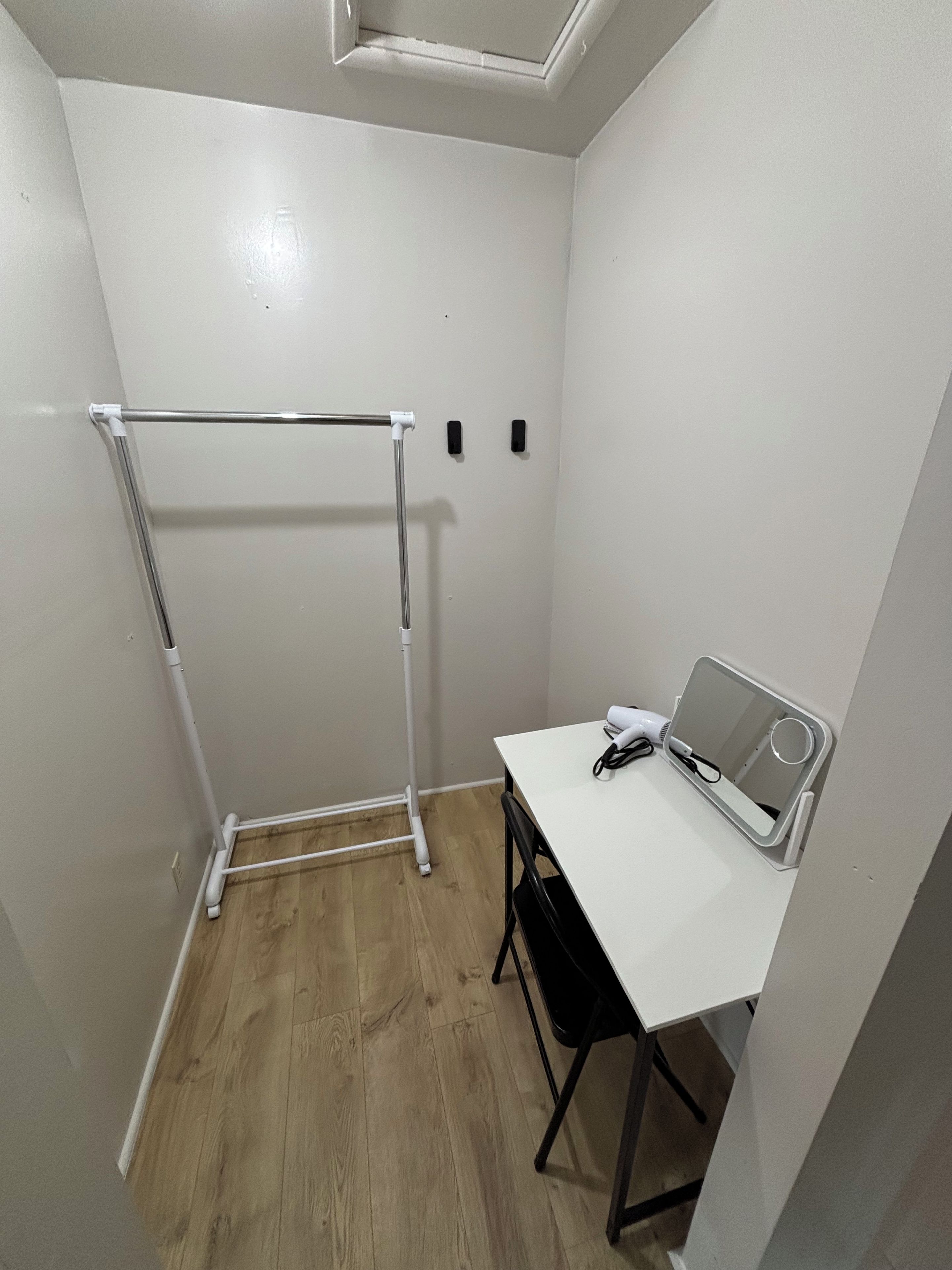 Une petite pièce avec un bureau blanc, une chaise, un radiateur blanc et une grille de ventilation blanche au plafond.