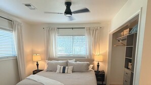 Un dormitorio con cama, mesitas de noche, un banco, ventilador de techo, ventana con cortinas y vestidor.