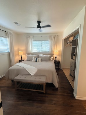 Ein Schlafzimmer mit einem Bett, Nachttischen, einer Bank, einem Deckenventilator, einem Fenster mit Vorhängen und einem begehbaren Kleiderschrank.