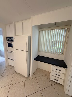 Cocina compacta con armarios blancos, horno integrado, refrigerador y ventana con persianas.