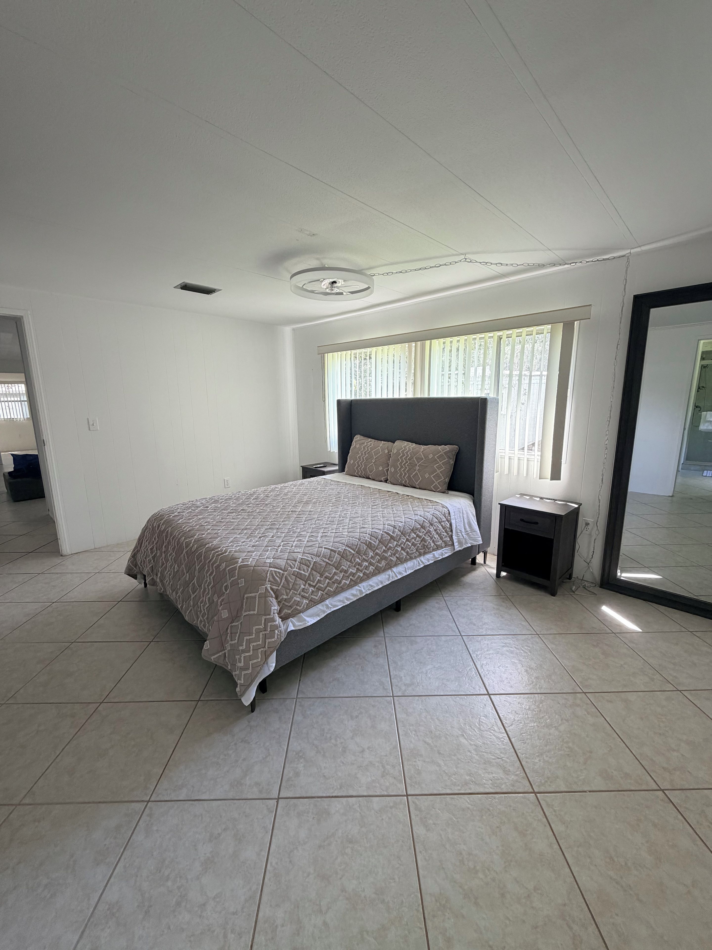 A bedroom with a bed, a nightstand, a mirror, and a window with blinds.