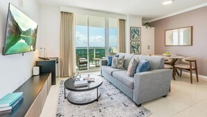 A modern living room with a grey sofa, a coffee table, and a TV mounted on the wall. There is a balcony with a view of the outdoors. - Bay View Beauty: 1/1.5 Bath Condo in Coconut Grove! Parking Included (Miami)