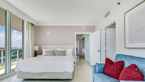 A modern hotel room with a large bed, a blue sofa with red cushions, and a city view through large windows. - Bay View Beauty: 1/1.5 Bath Condo in Coconut Grove! Parking Included (Miami)