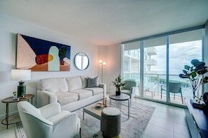 A modern living room with a sofa, coffee table, and a view of a body of water through large windows. - Direct Bayview One Bedroom Condo in Coconut Grove-includes Parking (Miami)
