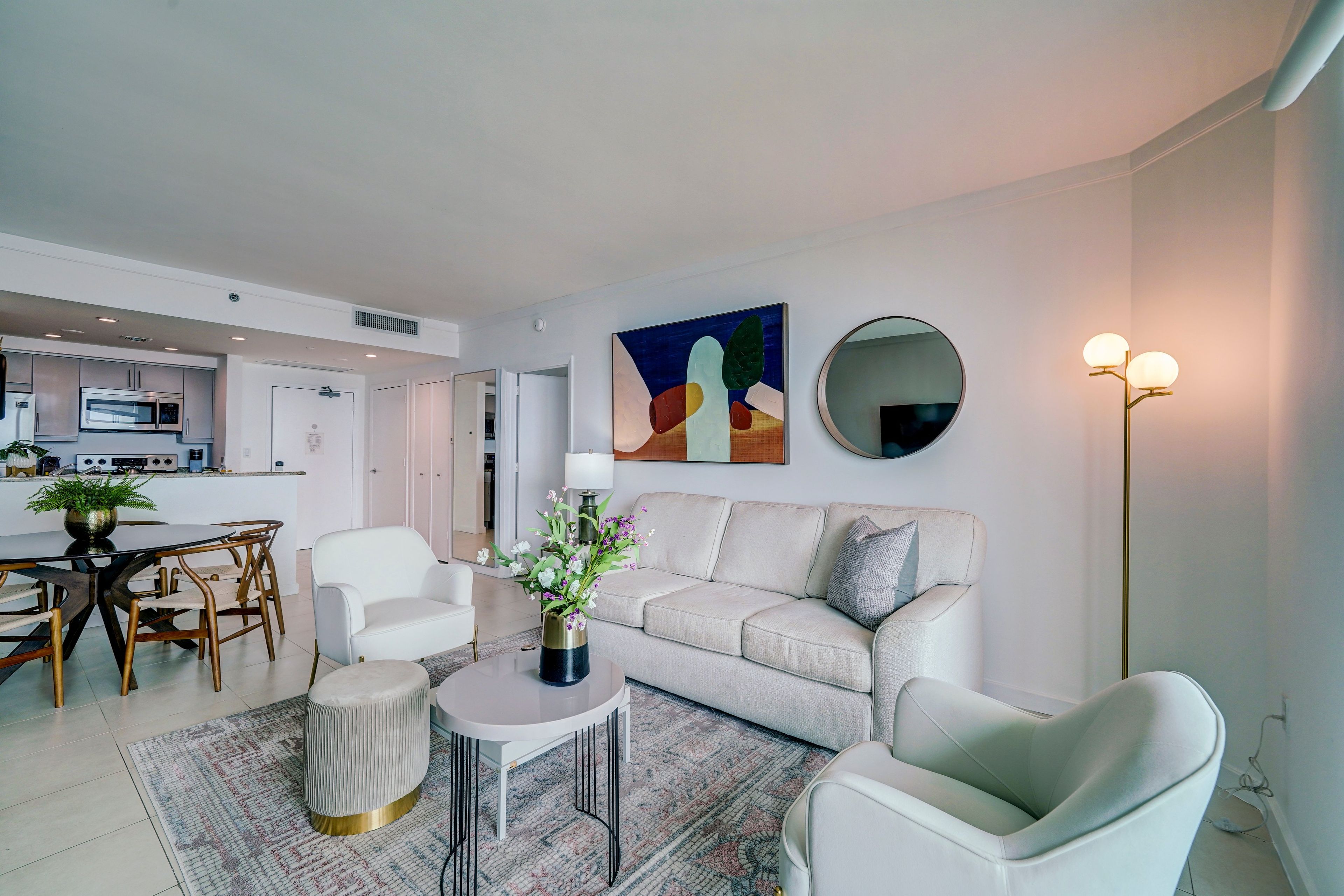 Een moderne woonkamer met een bank, stoelen, een salontafel en een eethoek met een tafel en stoelen. Achtergrond toont een keuken met een magnetron en kastjes.