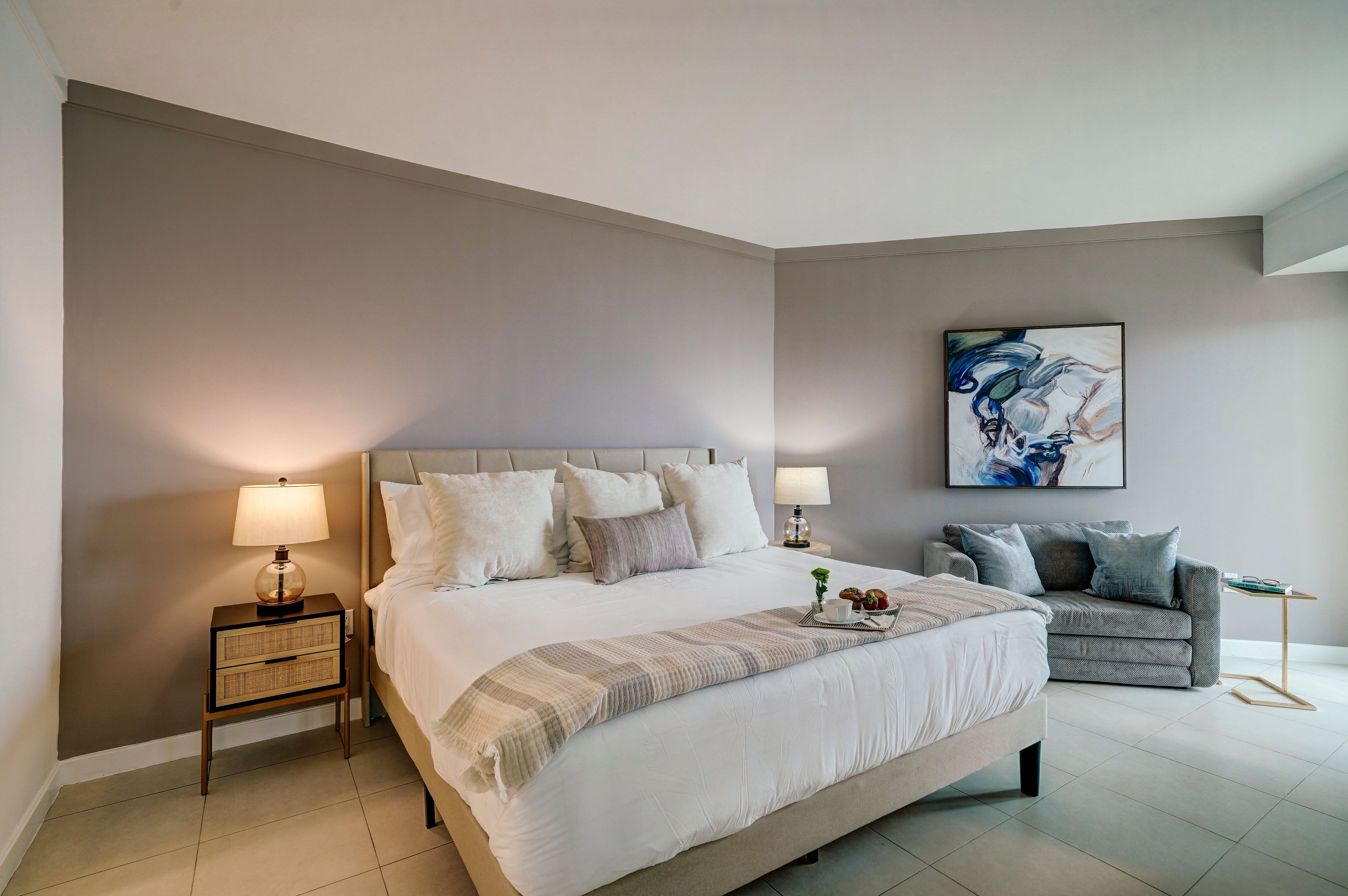 A hotel room with a large bed, bedside lamps, a side table, a sofa, and a painting on the wall.