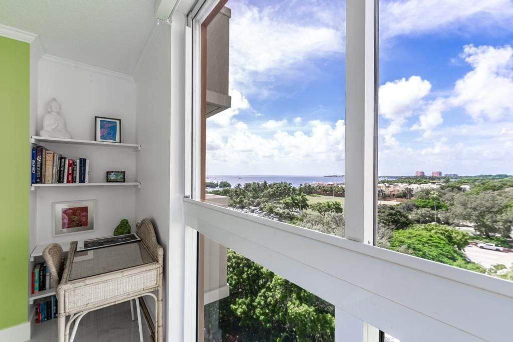 A room with a view of a coastal landscape, a white wall with a shelf holding books and a statue, and a wicker chair.