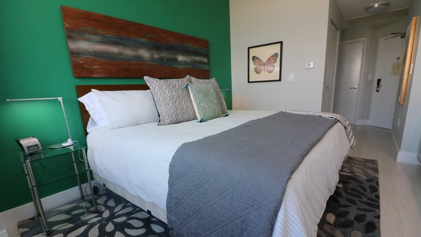 A neatly made bed with a patterned blanket and pillows, a bedside table with a lamp, and a butterfly wall art piece. - Renovated 21st Fl. Private Unit Located in Coconut Grove Hotel! (Miami)