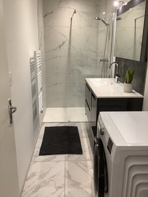 A modern bathroom with a white sink, a mirror, a shower, and a black rug. - Cozy apartment in downtown Marseillan – Sleeps 4 (Marseillan)