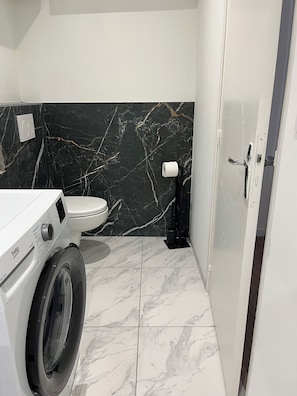 A compact bathroom with a washing machine, toilet, and a marble-tiled wall. - Cozy apartment in downtown Marseillan – Sleeps 4 (Marseillan)