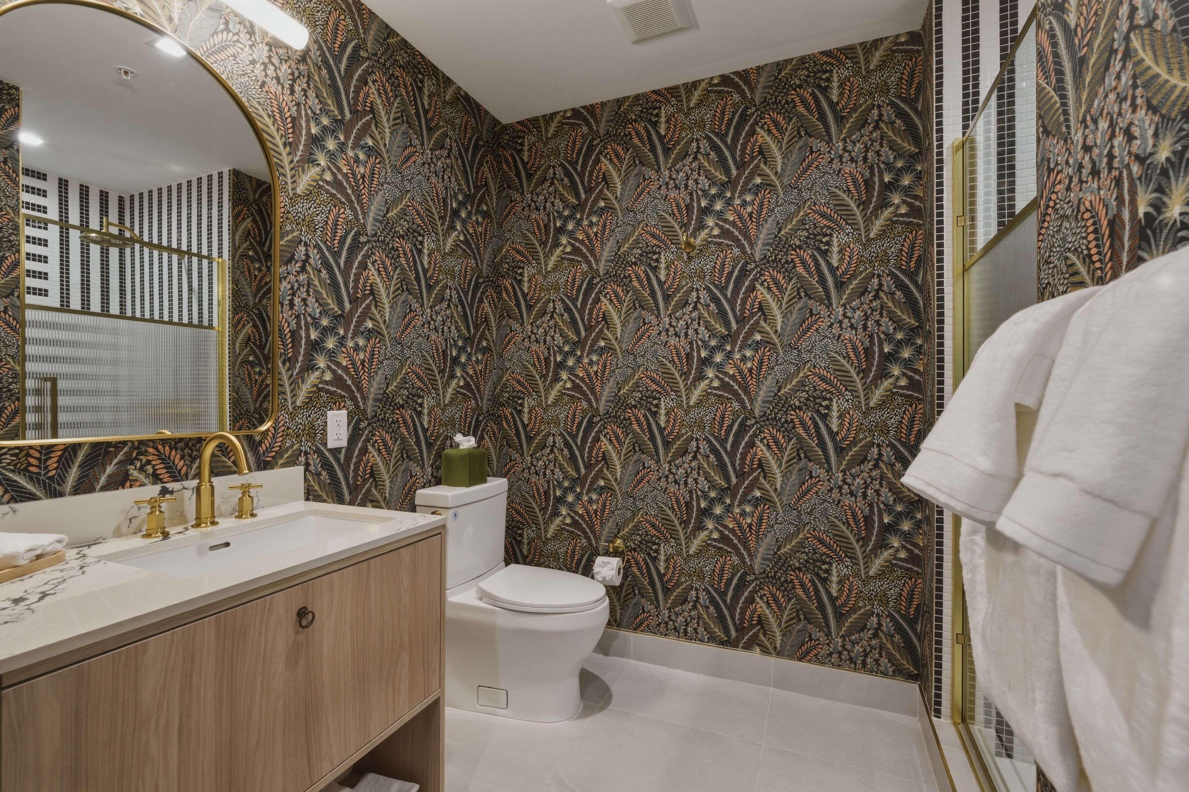 A bathroom with a large mirror, a sink with gold fixtures, a toilet, and a shower area.