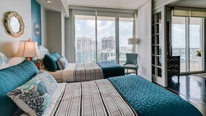 Une chambre moderne avec un grand lit, un fauteuil et une vue sur le paysage urbain grâce à de grandes fenêtres.