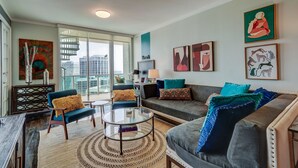 Un salon moderne avec une table basse en verre, un canapé et un fauteuil. Des œuvres d’art ornent les murs et de grandes fenêtres offrent une vue sur la ville.