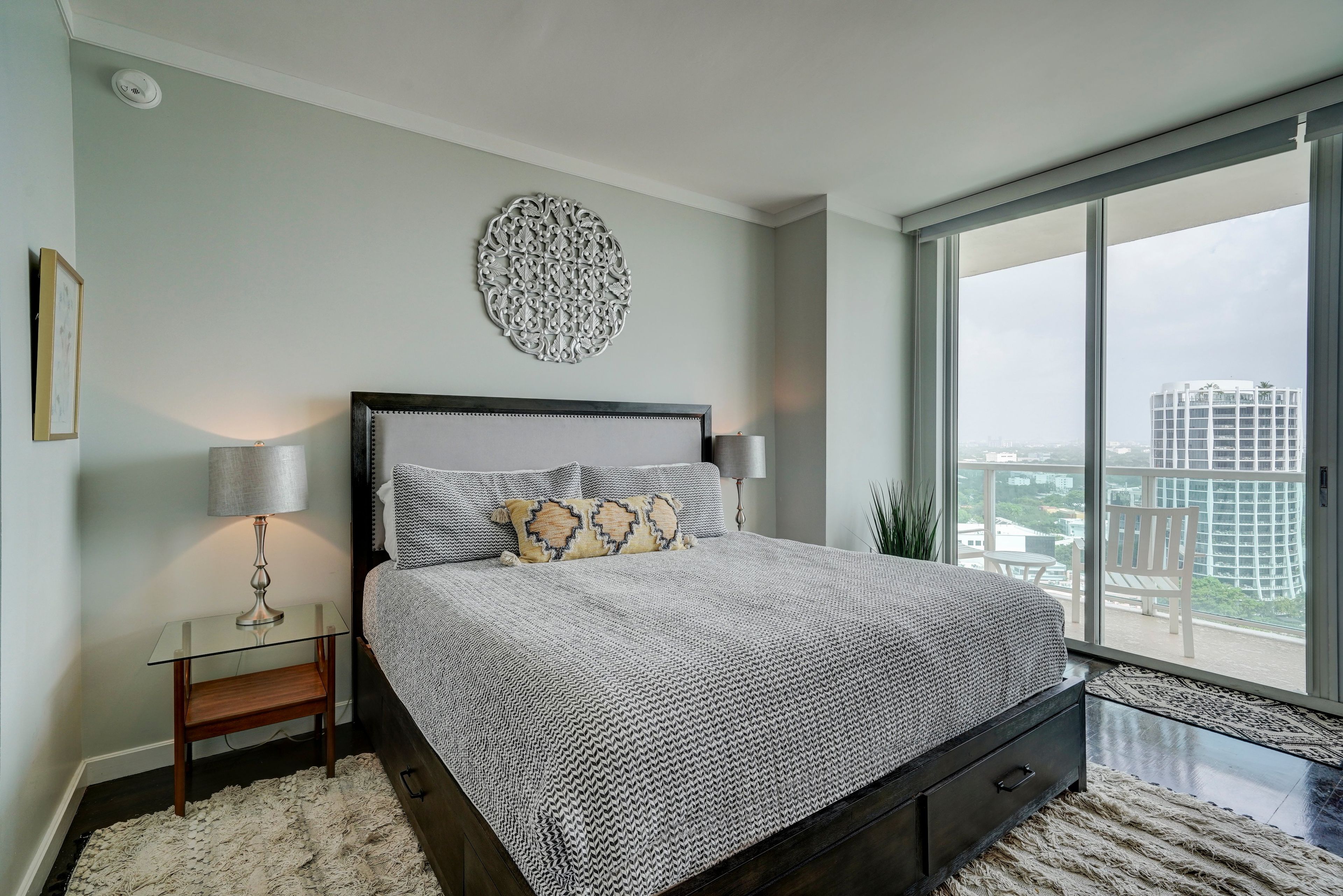 A bedroom with a large bed, a nightstand, a lamp, a decorative wall piece, and a view of a cityscape through a large window.