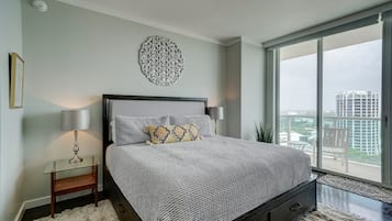 A bedroom with a large bed, a nightstand, a lamp, a decorative wall piece, and a view of a cityscape through a large window.