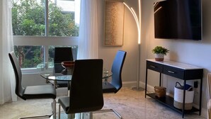 Een moderne woonkamer met een glazen eettafel, zwarte stoelen, een zwarte dressoir met een televisie, en een groot raam met gordijnen.