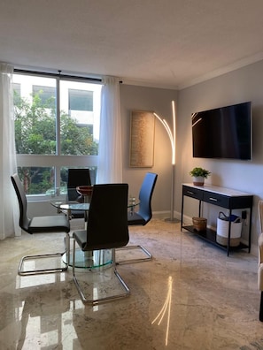 A modern living room with a glass dining table, black chairs, a black console table with a TV, and a large window with curtains. - Cozy 1 Bedroom Unit Located in a Condo/hotel in the Heart of Coconut Grove-free Parking (Miami)