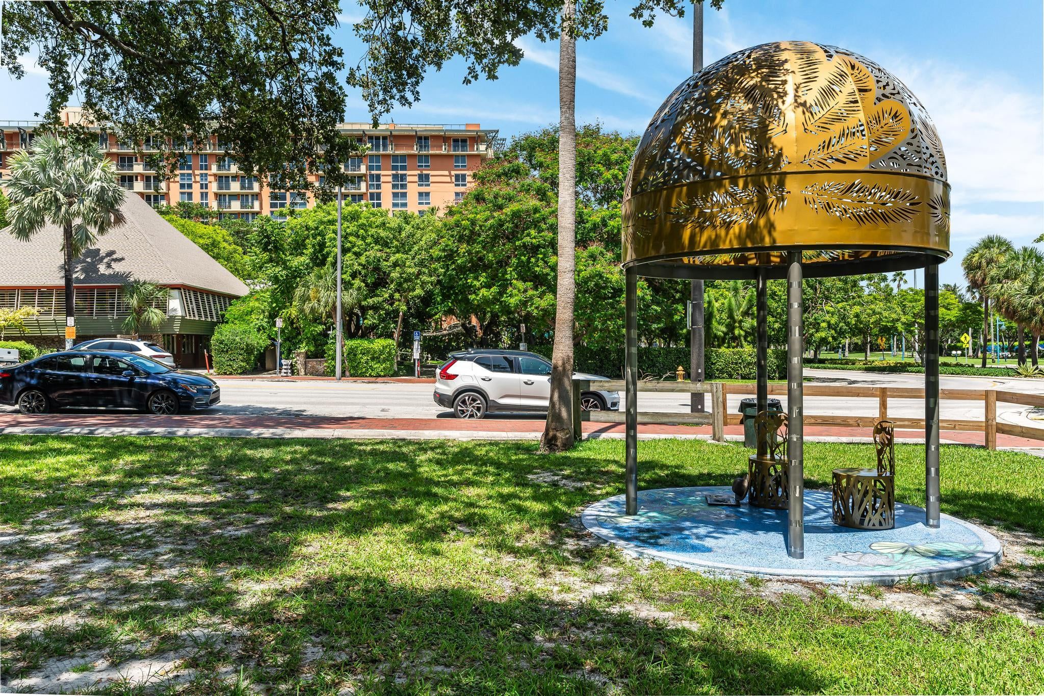Ein Pavillon mit goldenem Blattmuster, umgeben von Gras und Bäumen, mit einem Gebäude und geparkten Autos im Hintergrund.