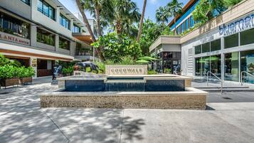 Un espace extérieur moderne doté d’une piscine, de palmiers et de boutiques, notamment un bar branché et un magasin de cosmétiques.