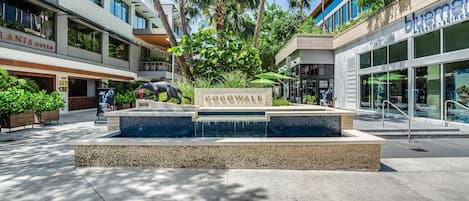 Un espacio exterior moderno con piscina, palmeras y locales comerciales, incluyendo un club nocturno y una tienda de maquillaje.