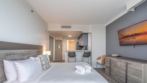 Ett hotellrum med en säng, ett skrivbord, en stol, en tv och ett litet köksutrymme i bakgrunden.