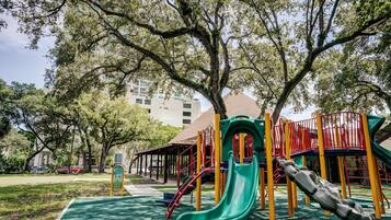 Un parque infantil con un tobogán verde y estructuras de juego amarillas y rojas, rodeado de árboles y un edificio al fondo.
