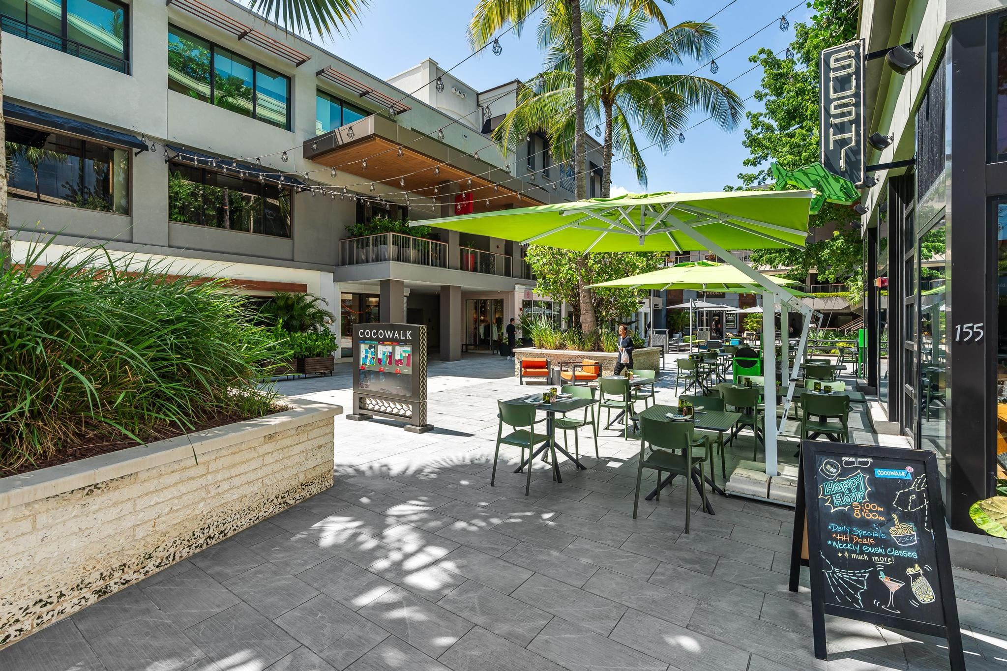 Un área de café al aire libre con mesas y sillas, un cartel con las ofertas especiales y un edificio con el letrero 'Cocowalk'.