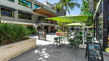 Un área de café al aire libre con mesas y sillas, un cartel con las ofertas especiales y un edificio con el letrero 'Cocowalk'.
