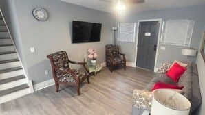 Sala de estar com escada, dois poltronas, mesa de centro de vidro, sofá, televisão e porta com placa.